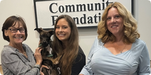 Women smiling with a small dog, standing in front of a foundation sign.